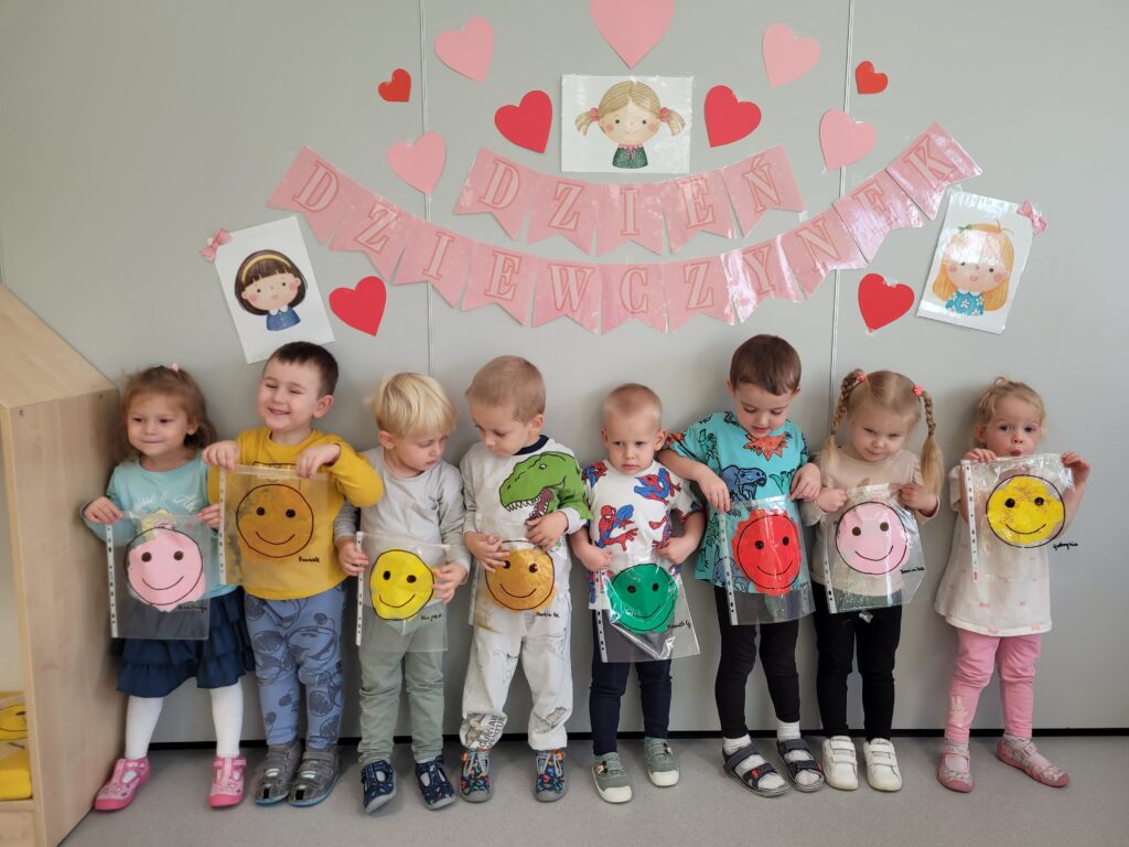 Trzy dziewczynki i pięciu chłopców stoi na tle dekoracji z okazji Dnia dziewczynki. Wszyscy trzymają w ręce symbol uśmiechniętej buźki.