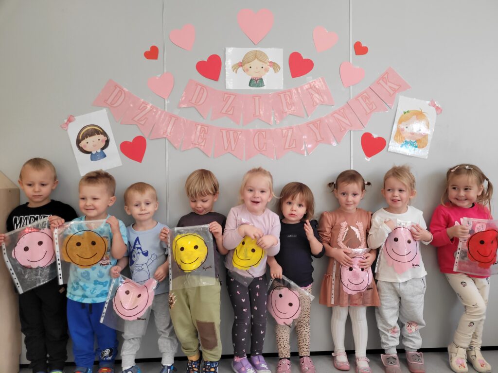 Pięć dziewczynek i czterech chłopców stoi na tle dekoracji z okazji Dnia dziewczynki. Wszyscy trzymają w ręce symbol uśmiechniętej buźki.