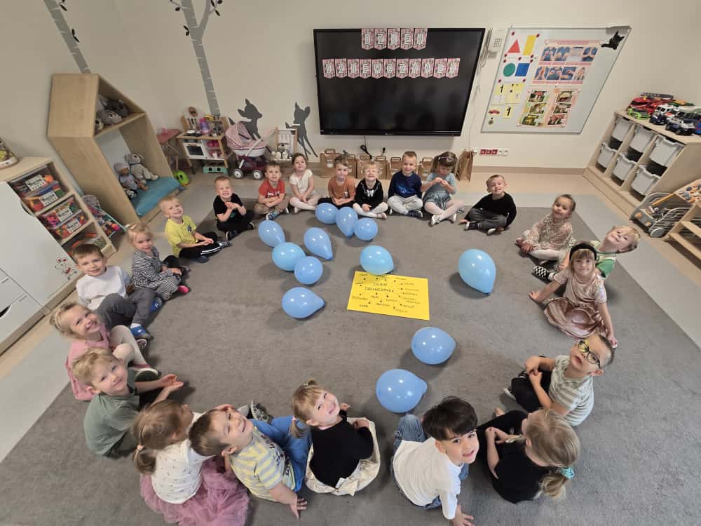 Duża grupa dzieci siedzi na szarym dywanie w kole, w środku niebieskie balony i żółta kartka