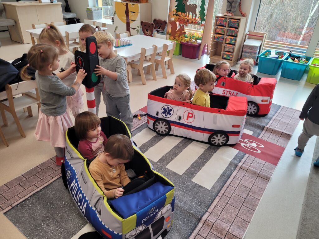 Dzieci dwójkami siedzą w samochodach (policji, karetki, straży pożarnej). czwórka dzieci ogląda sygnalizator świetlny.
