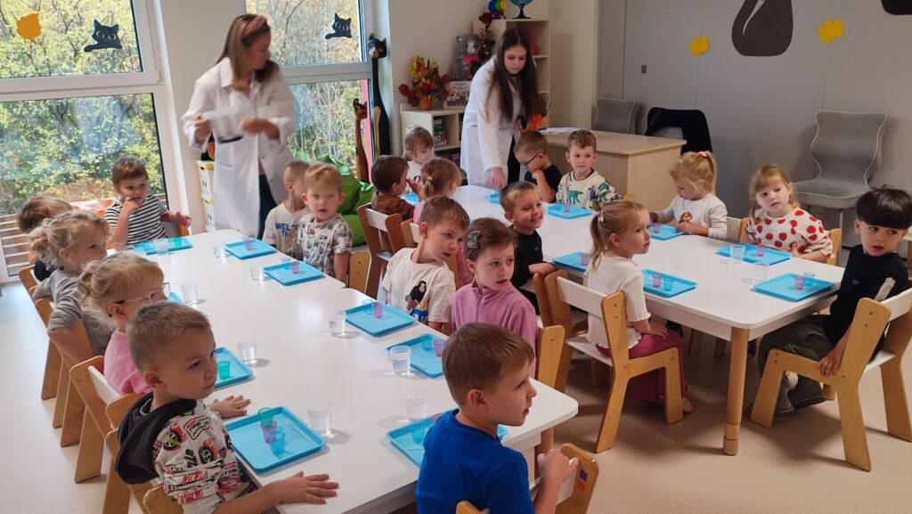 Duża grupa dzieci siedzi przy długim białym stole, na których leżą niebieskie tacki.