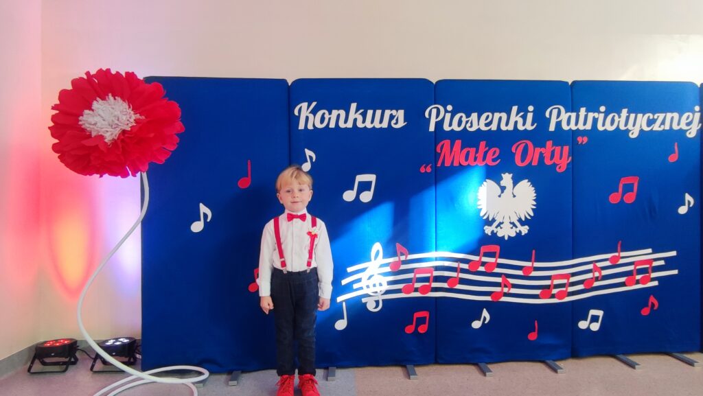 Chłopiec ubrany w białą koszulę, czerwoną muszkę i czerwone szelki pozuje do zdjęcia. Za nim duża tablica niebieska z godłem Polski, nutami i napisem. To wszystko w kolorach białych i czerwonych.