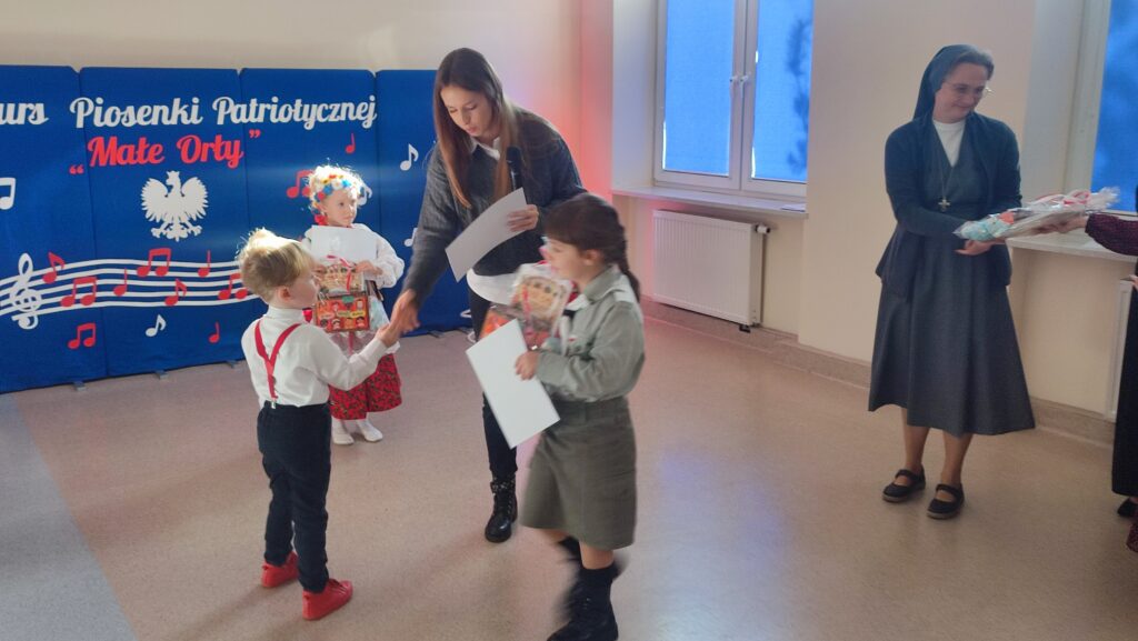 Chłopiec ubrany w białą koszulę, czerwoną muszkę i czerwone szelki odbiera nagrodę i dyplom z rąk kobiety.