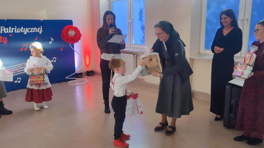 Chłopiec ubrany w białą koszulę, czerwoną muszkę i czerwone szelki odbiera nagrodę i dyplom z rąk siostry Salezianki.