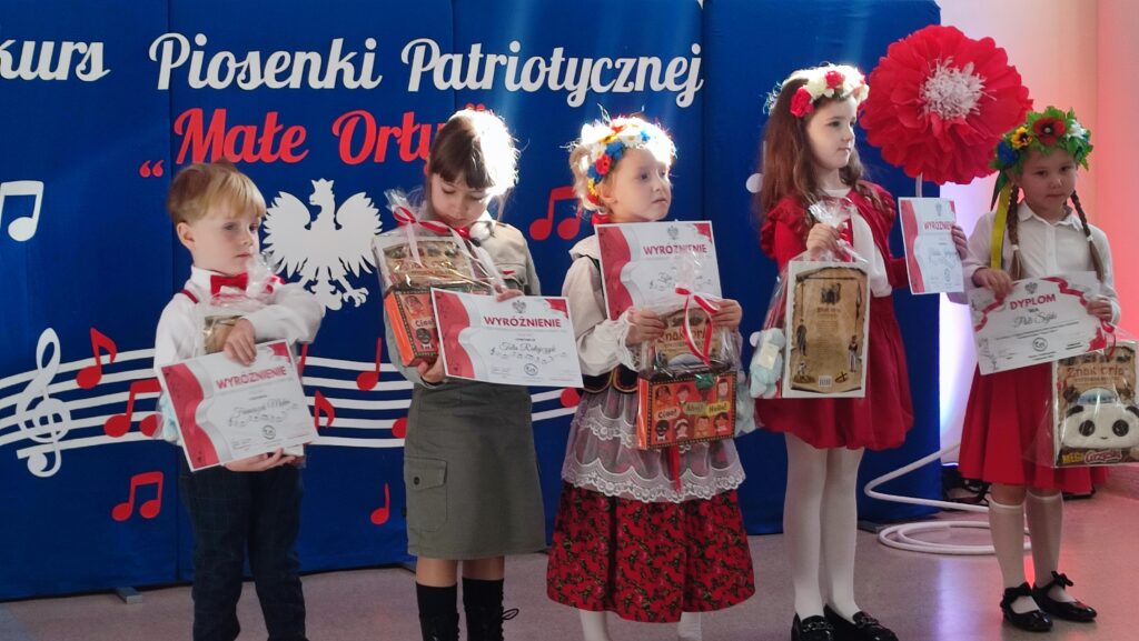 Chłopiec ubrany w białą koszulę, czerwoną muszkę i czerwone szelki pozuje do wspólnego zdjęcia. Za nim duża tablica niebieska z godłem Polski, nutami i napisem. To wszystko w kolorach białych i czerwonych. Obok chłopca stoją dziewczynki. Wszyscy trzymają dyplomy i nagrody.