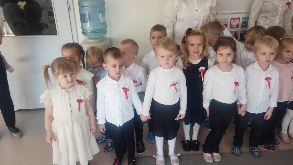 Bardzo duża grupa dzieci ubrana w stroje biało czarne w wpiętymi kotylionami narodowymi stoi na szarym dywanie i śpiewa hymn Polski.