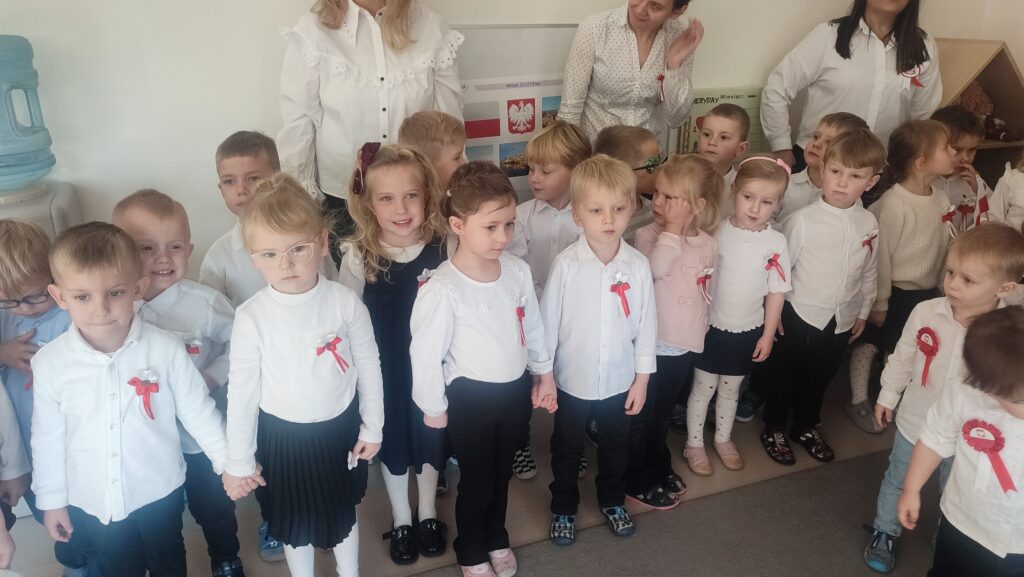 Bardzo duża grupa dzieci ubrana w stroje biało czarne w wpiętymi kotylionami narodowymi stoi na szarym dywanie i śpiewa hymn Polski.
