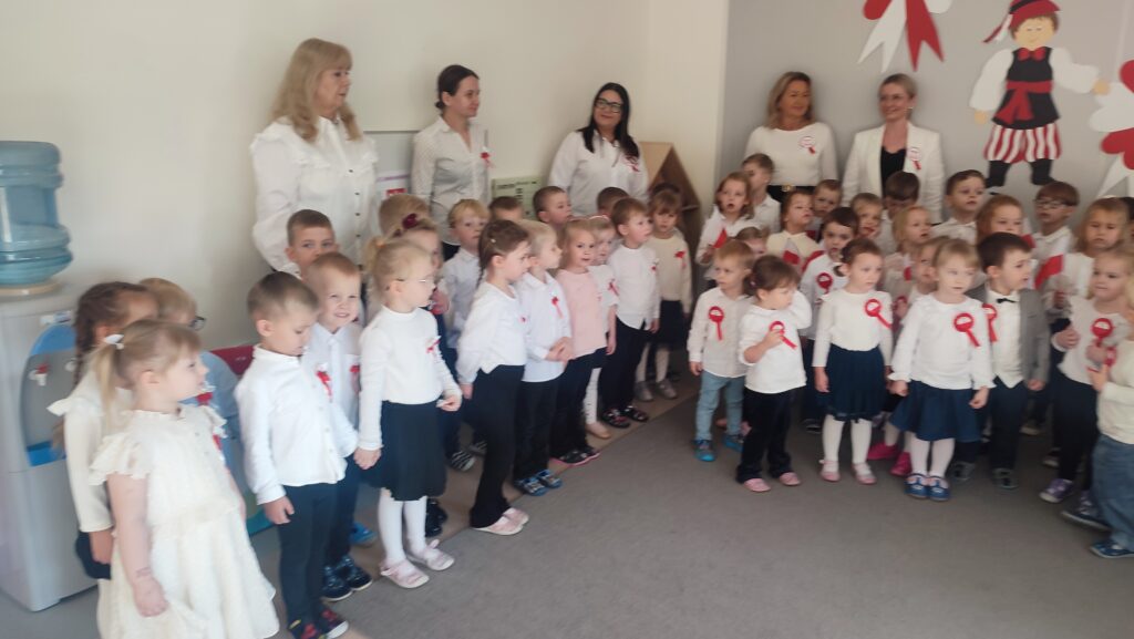 Bardzo duża grupa dzieci ubrana w stroje biało czarne w wpiętymi kotylionami narodowymi stoi na szarym dywanie i śpiewa hymn Polski.