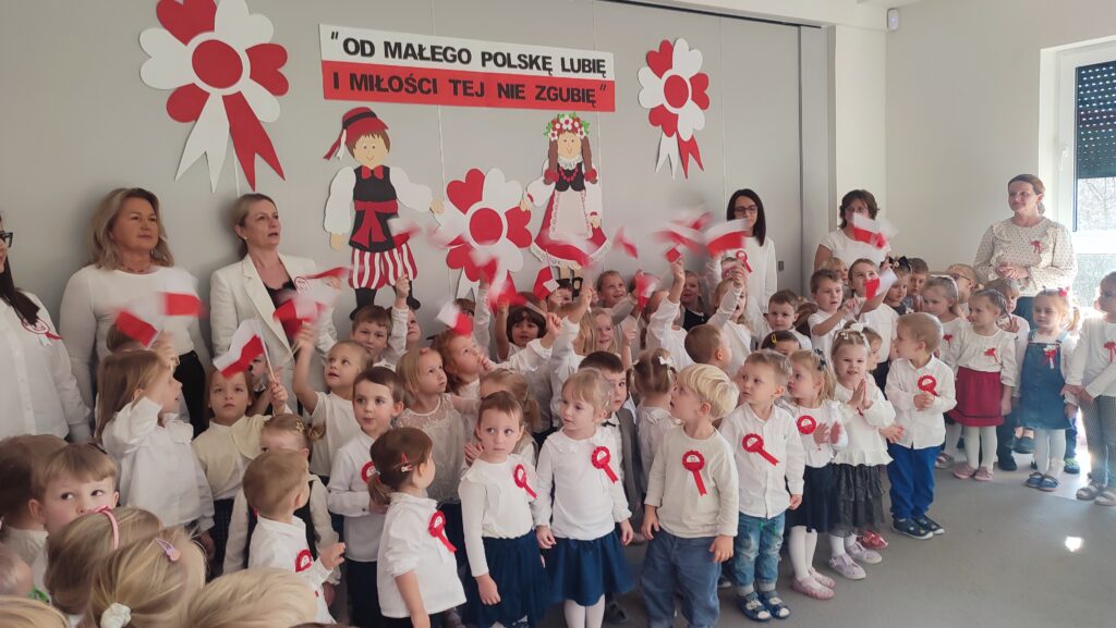 Bardzo duża grupa dzieci ubrana w stroje biało czarne w wpiętymi kotylionami narodowymi stoi na szarym dywanie i śpiewa hymn Polski. Za nimi szara ściana z dekoracją w barwach biało-czerwonych.