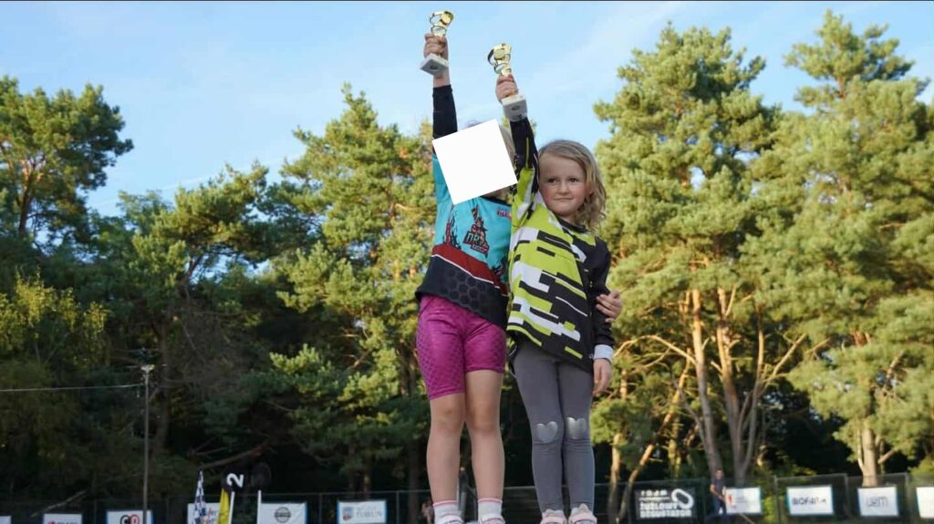 dwie dziewczynki stoją na podium z pucharami wysoko podniesionymi 