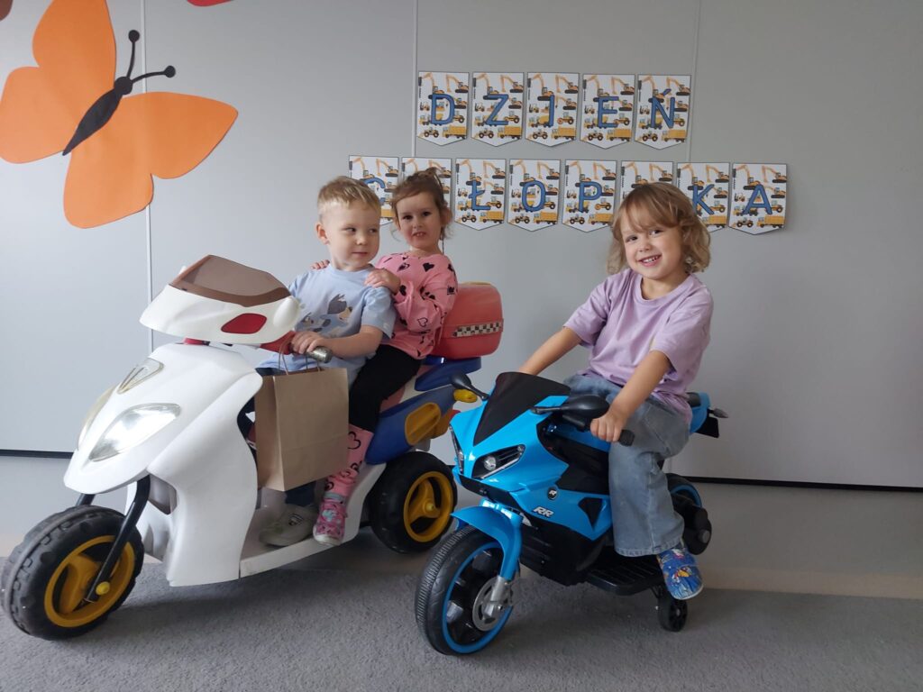 Chłopiec z dziewczynką w bluzce w serduszka siedzą na białym motorze oraz chłopiec w fioletowej bluzce siedzi na niebieskim motorze