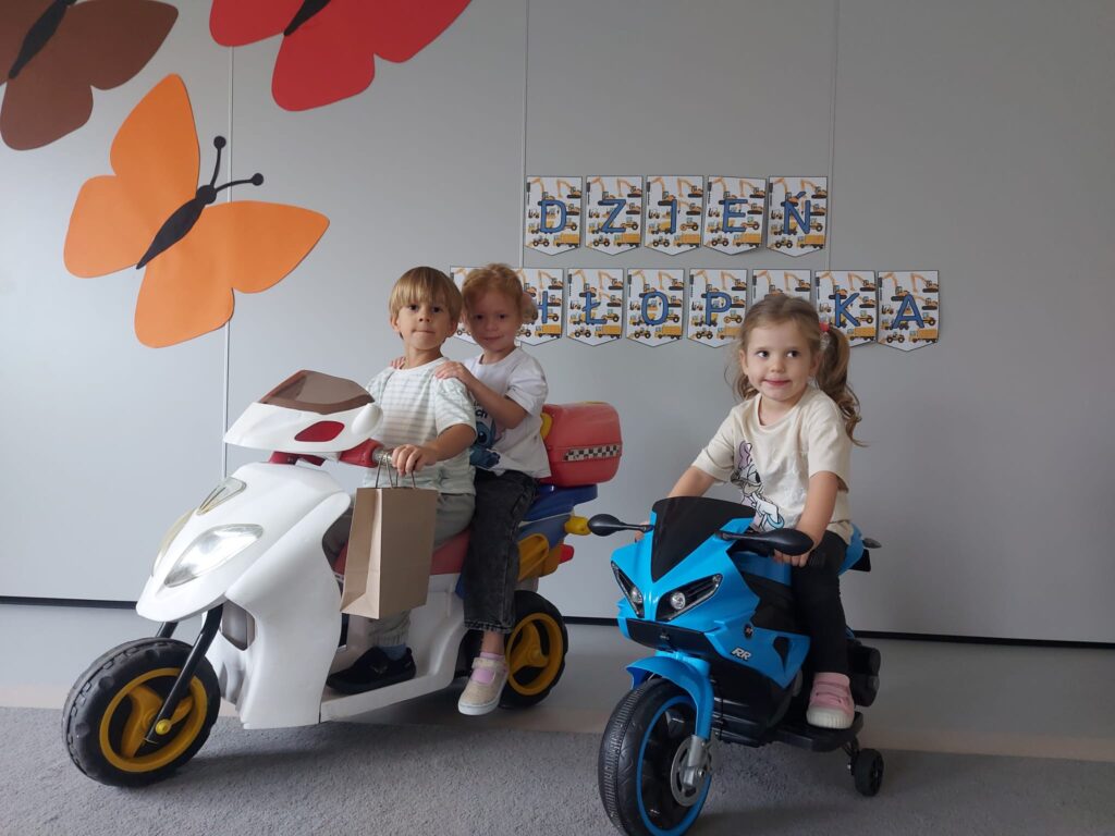 chłopiec z dziewczynką siedzą na białym zabawkowym motorze oraz dziewczynka w kitkach siedzi na niebieskim motorze