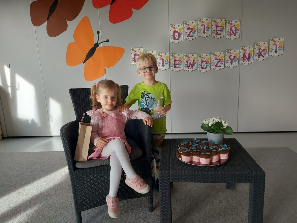 dziewczynka siedzi na wiklinowym fotelu, chłopiec stoi za nią, na stoliku stoją kwiatki i babeczki na talerzu