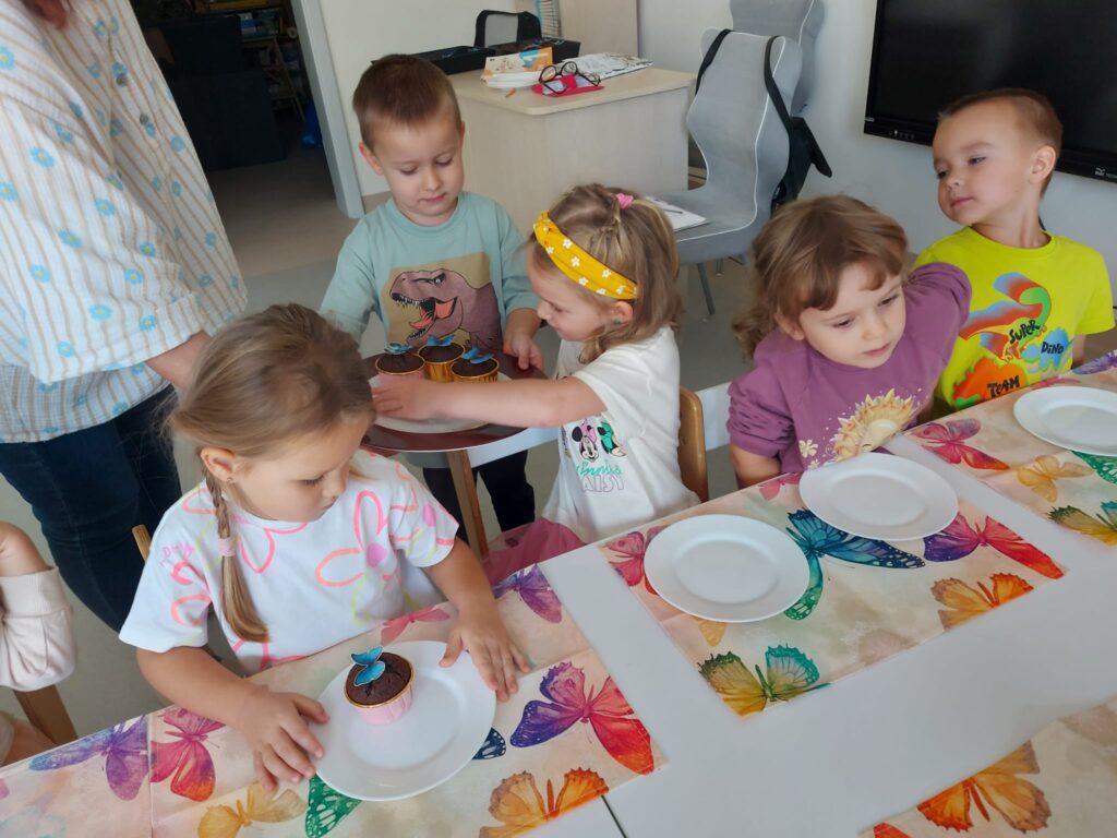 dziewczynki siedzą przy stoliku, chłopiec trzyma talerz z babeczkami i częstuje nimi dzieci 