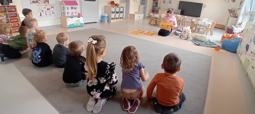 dzieci siedzą na dywanie pani pokazuje materiały edukacyjne, pies leży na legowisku