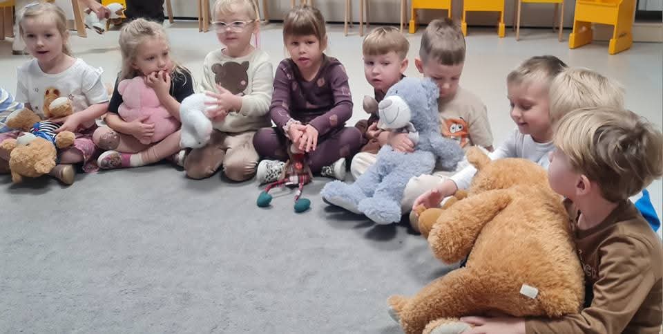 Dzieci siedzą na szarym dywanie w rękach trzymają pluszowe misie.