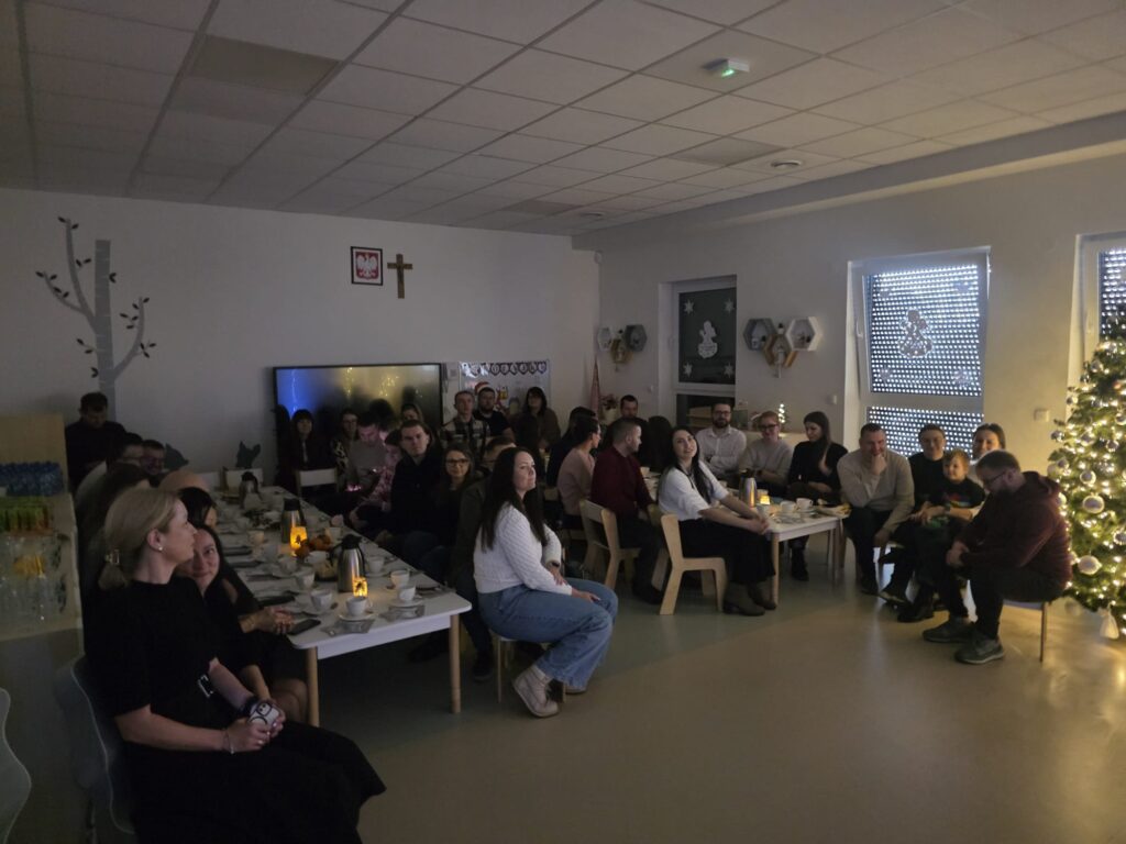 Duża grupa dorosłych ludzi siedzi na białych krzesłach przy świątecznym stole.