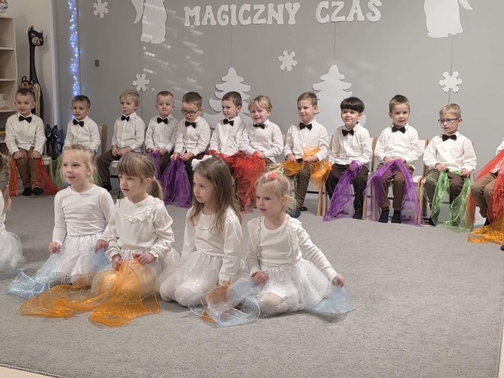 Duża grupa chłopców siedzi na białych krzesłach, w rękach trzymają kolorowe chustki. Przed nimi siedzą dziewczynki ubrane na biało, w rękach trzymają kolorowe chustki.