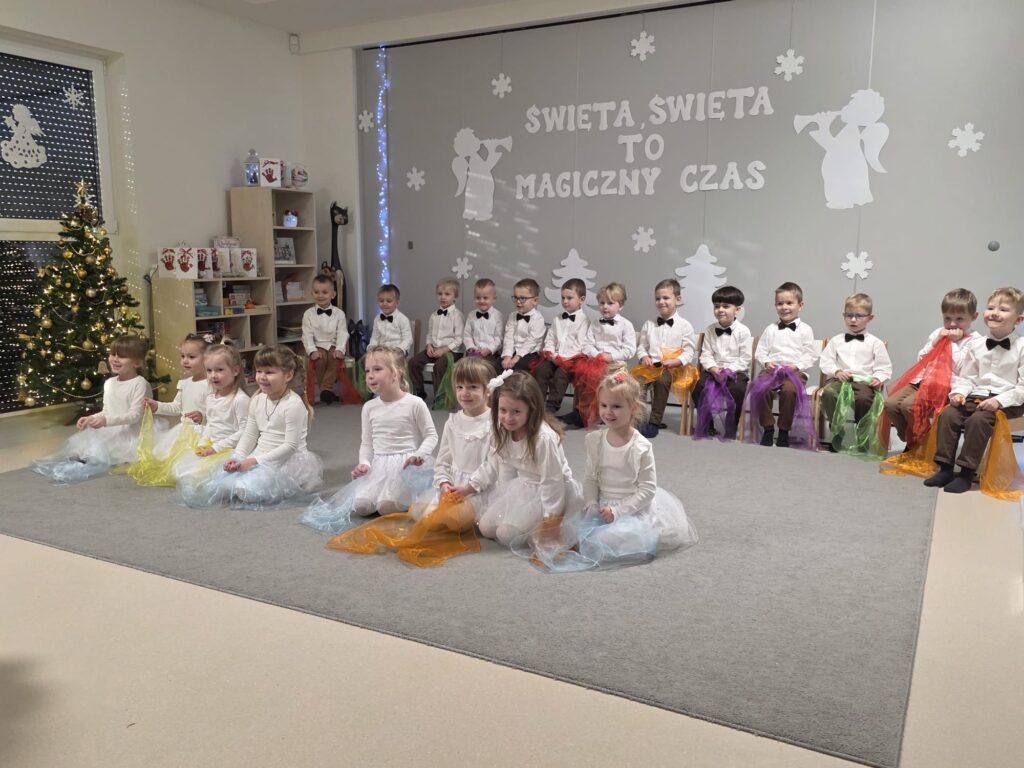 Duża grupa chłopców siedzi na białych krzesłach, w rękach trzymają kolorowe chustki. Przed nimi siedzą dziewczynki ubrane na biało, w rękach trzymają kolorowe chustki.