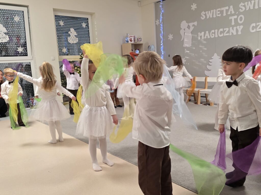 Duża grupa dzieci ubranych na biało, brązowo trzymają w rękach kolorowe chustki i tańczą na szarym dywanie.