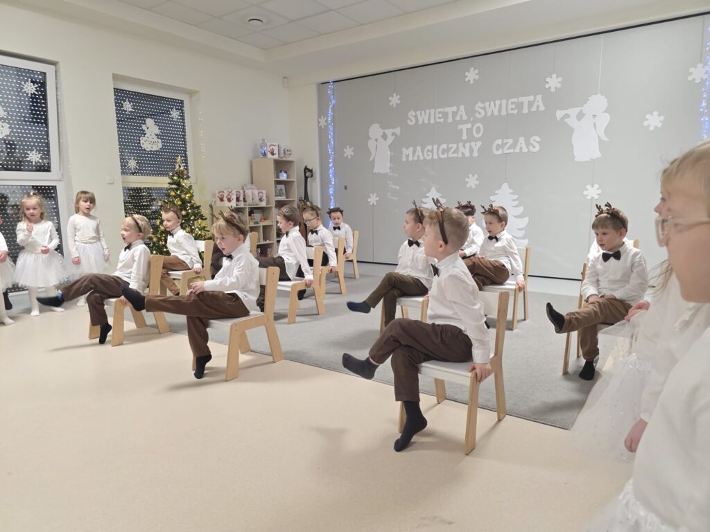Chłopcy ubrani w brązowe spodnie, w białe koszule z opaską renifera na głowie tańczą na białych krzesłach na szarym dywanie.
