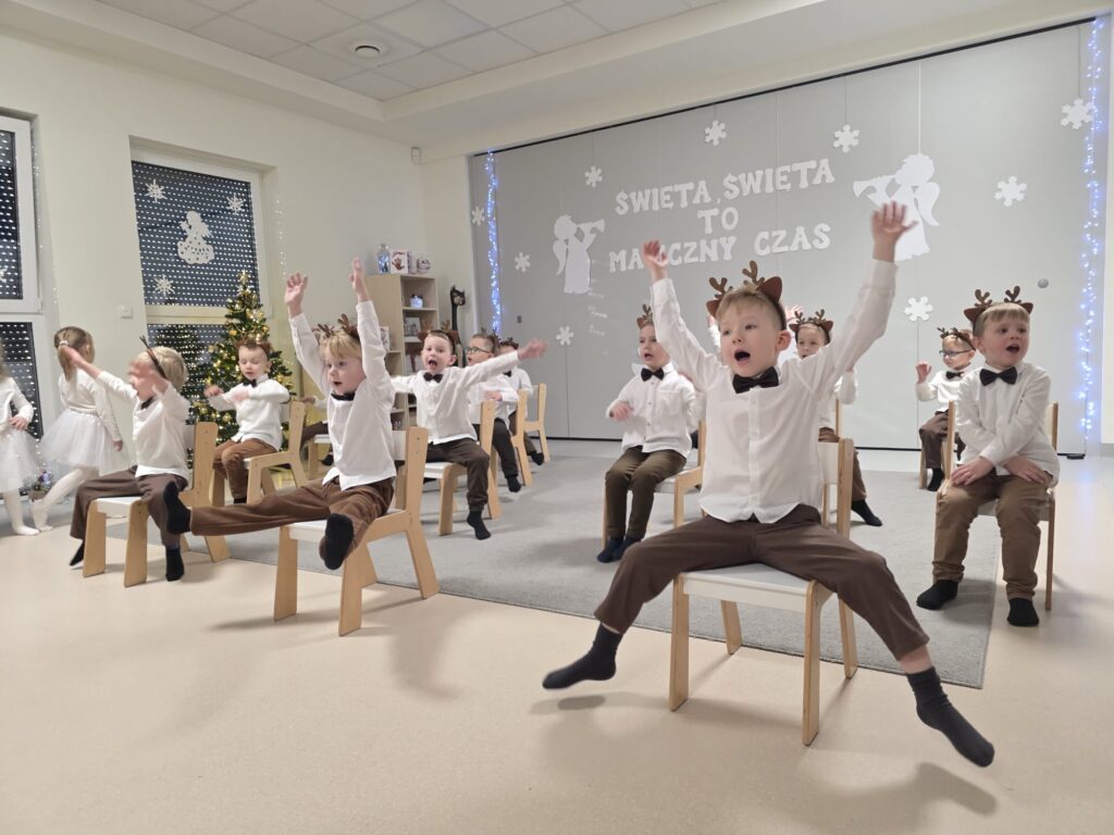 Chłopcy ubrani w brązowe spodnie, w białe koszule z opaską renifera na głowie tańczą na białych krzesłach na szarym dywanie.