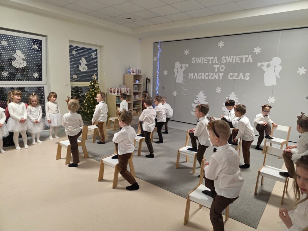 Chłopcy ubrani w brązowe spodnie, w białe koszule z opaską renifera na głowie tańczą na białych krzesłach na szarym dywanie.