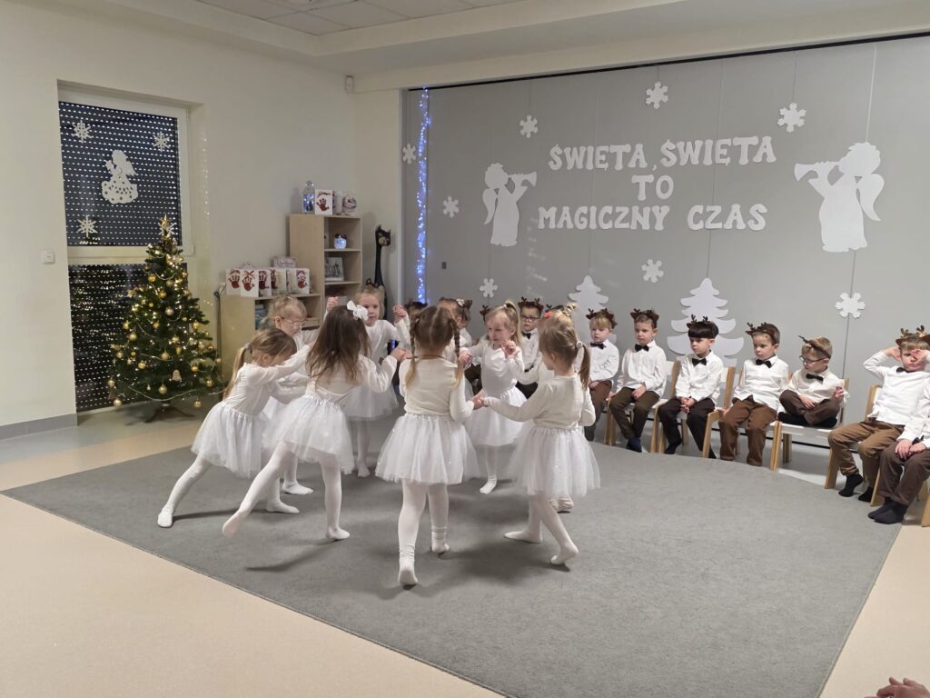 Dziewczynki ubrane na biało tańczą na szarym dywanie.