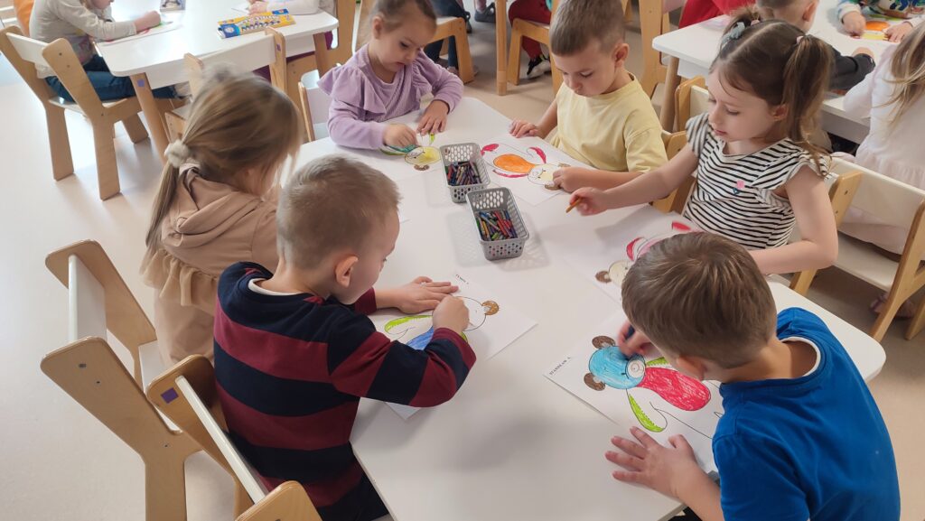 Dzieci siedzą przy białych stolikach i kolorują kredkami postać misia.