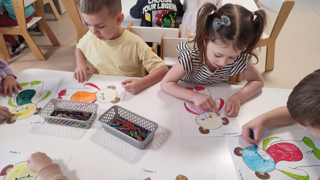 Dzieci siedzą przy białych stolikach i kolorują kredkami postać misia.