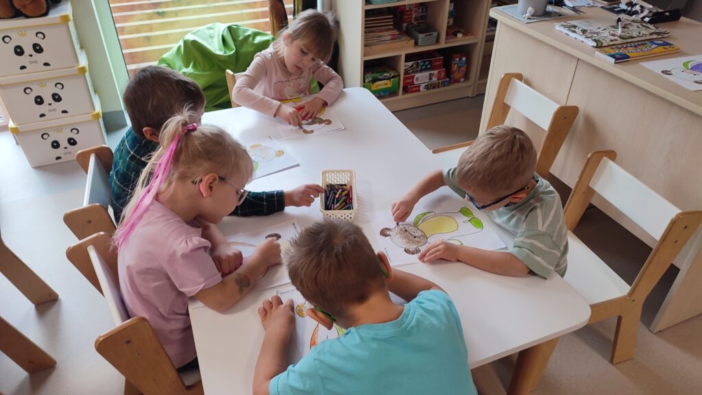 Dzieci siedzą przy białych stolikach i kolorują kredkami postać misia.