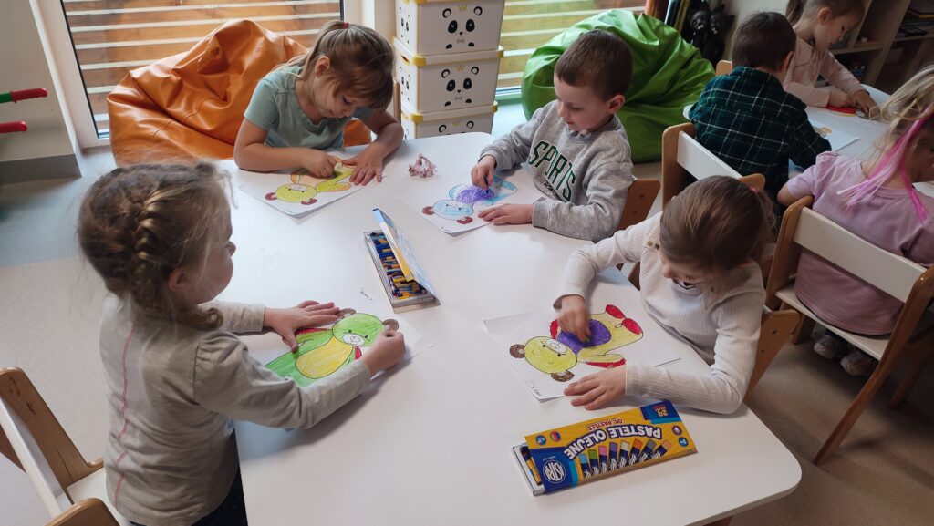 Dzieci siedzą przy białych stolikach i kolorują kredkami postać misia.