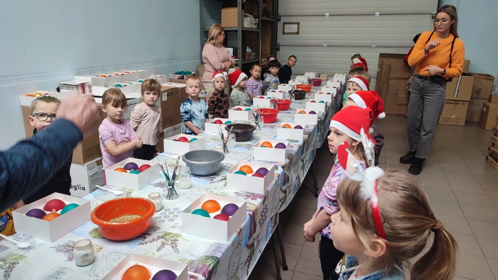 Dzieci stoją po obu stronach długiego stołu. Na stole kartony z bombkami, kolorowe miski z brokatem, pędzelki w słoikach. Niektóre dzieci mają na głowie czerwone czapki Mikołaja.