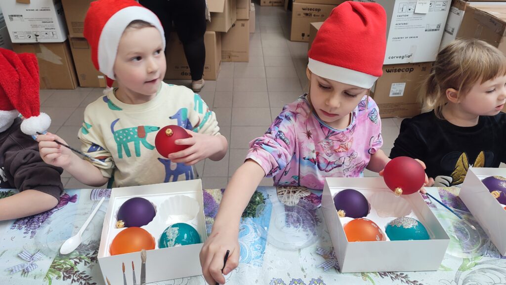 Dzieci w czerwonych czapkach Mikołaja stoją za stołem. W rękach trzymają bombkę i pędzel do malowania. Na stole stoją miski kolorowe z brokatem i pudełka z bombkami i pudełko z bombkami.