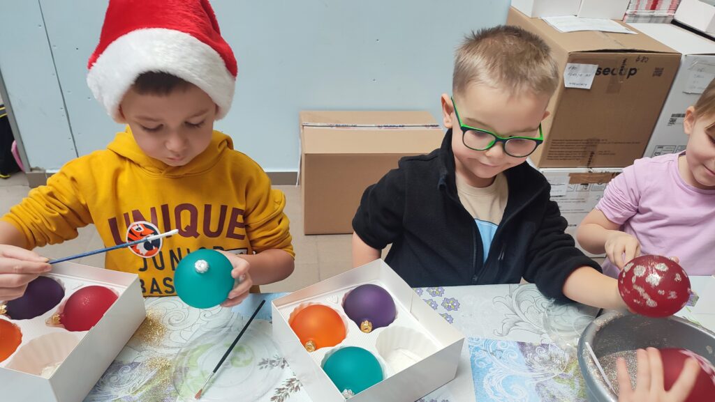 Dzieci w czerwonych czapkach Mikołaja stoją za stołem. W rękach trzymają bombkę i pędzel do malowania. Na stole stoją miski kolorowe z brokatem i pudełko z bombkami.