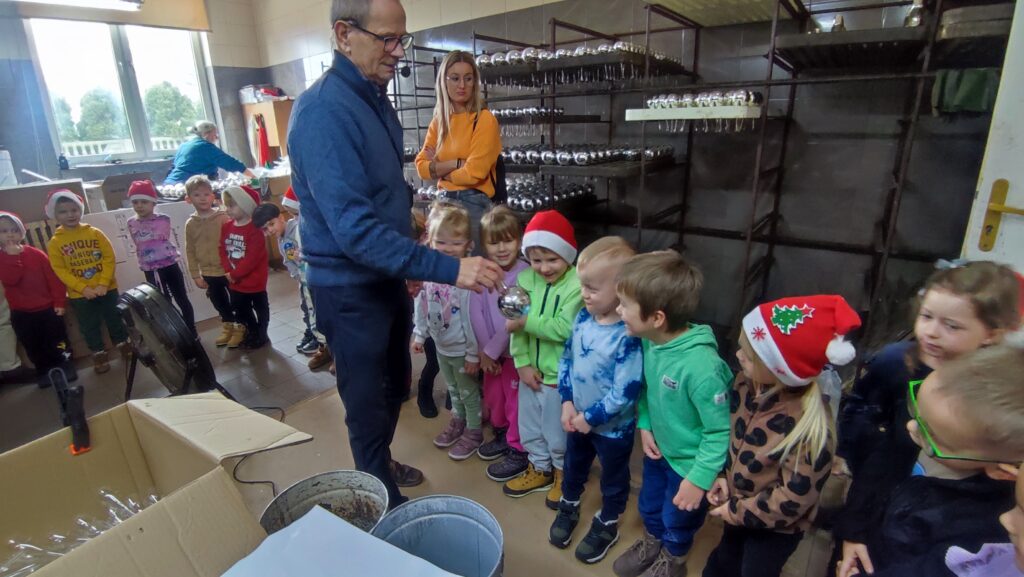Grupa dzieci ogląda srebrną bombkę pokazywaną przez mężczyznę. Za nimi regał ze srebrnymi bombkami.