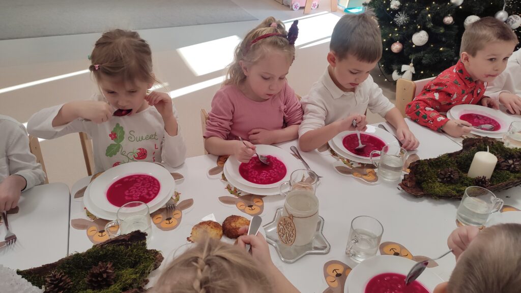 Duża grupa dzieci ubranych elegancko siedzi przy białych stołach, na których znajdują się potrawy wigilijne. Dzieci jedzą czerwony barszcz, rybę.