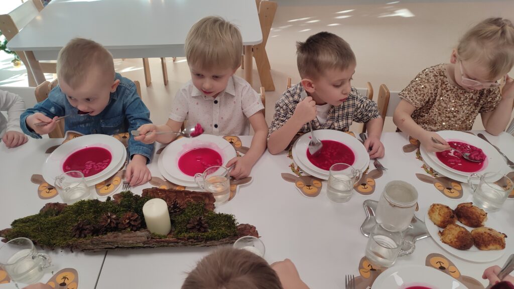 Duża grupa dzieci ubranych elegancko siedzi przy białych stołach, na których znajdują się potrawy wigilijne. Dzieci jedzą czerwony barszcz, rybę.