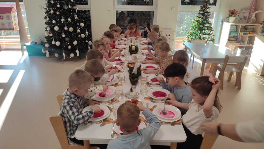 Duża grupa dzieci ubranych elegancko siedzi przy białych stołach, na których znajdują się potrawy wigilijne. Dzieci jedzą czerwony barszcz, rybę.