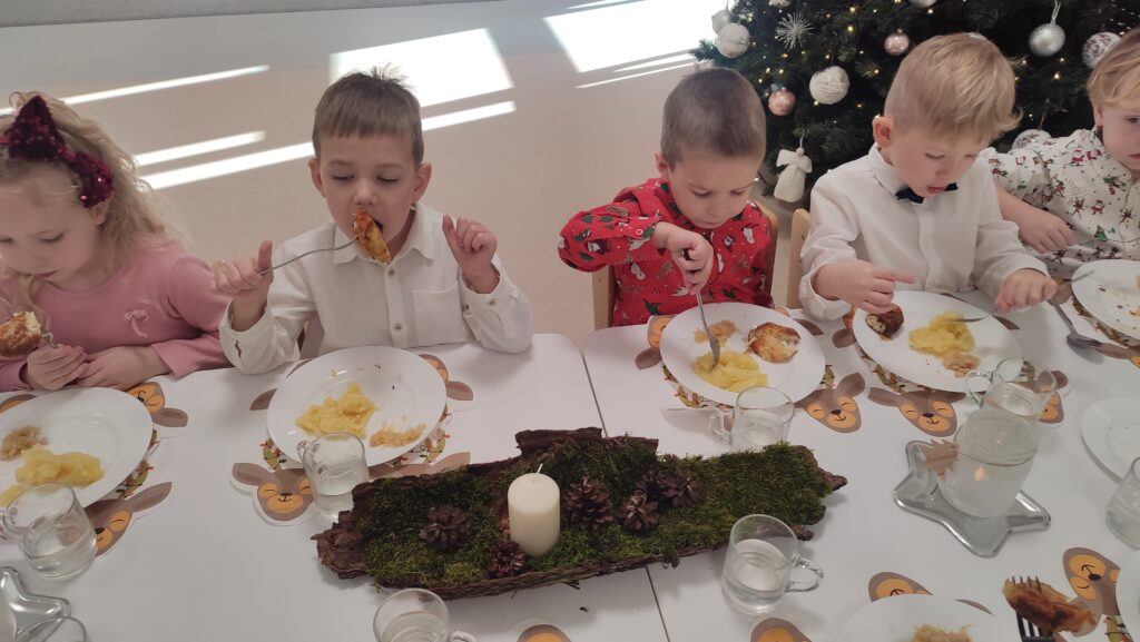 Duża grupa dzieci ubranych elegancko siedzi przy białych stołach, na których znajdują się potrawy wigilijne. Dzieci jedzą czerwony barszcz, rybę.