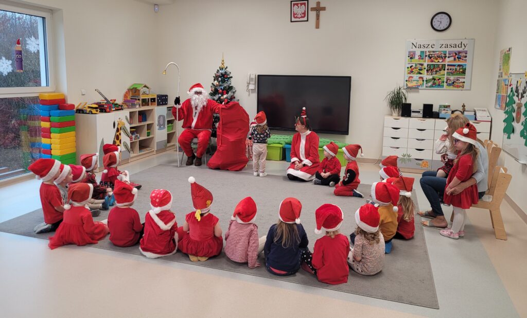 Dzieci i Pani Mikołajowa siedzą w kole na dywanie, obok siedzi Pani przy, której stoi dziewczynka. Mikołaj siedzi na krześle i trzyma worek z prezentami.