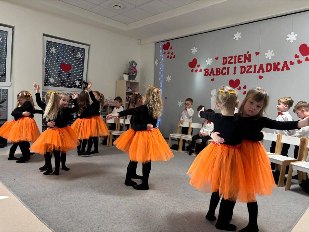 Grupa dziewczynek ubrane na czarno w pomarańczowe spódniczki tańczą na szarym dywanie.