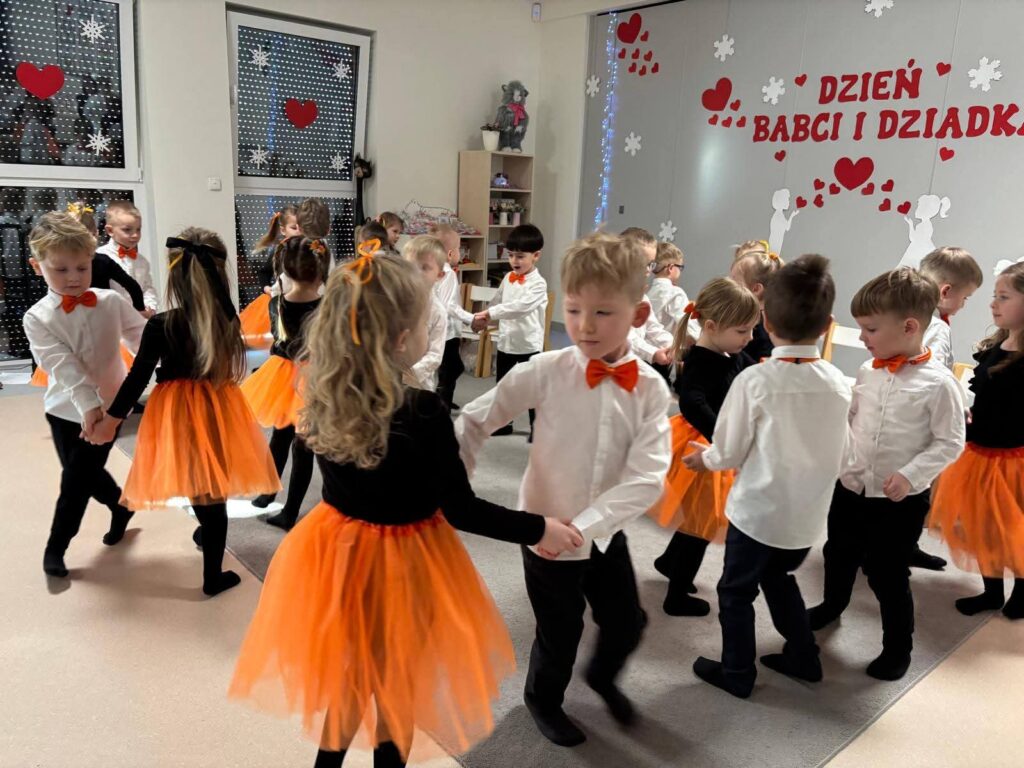 Chłopcy i dziewczynki ubrani w biało-czarno-pomarańczowe stroje, stoją na szarym dywanie i tańczą do muzyki.