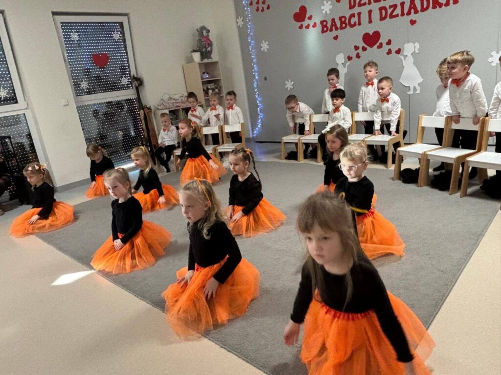Grupa dziewczynek ubrane na czarno w pomarańczowe spódniczki tańczą na szarym dywanie.