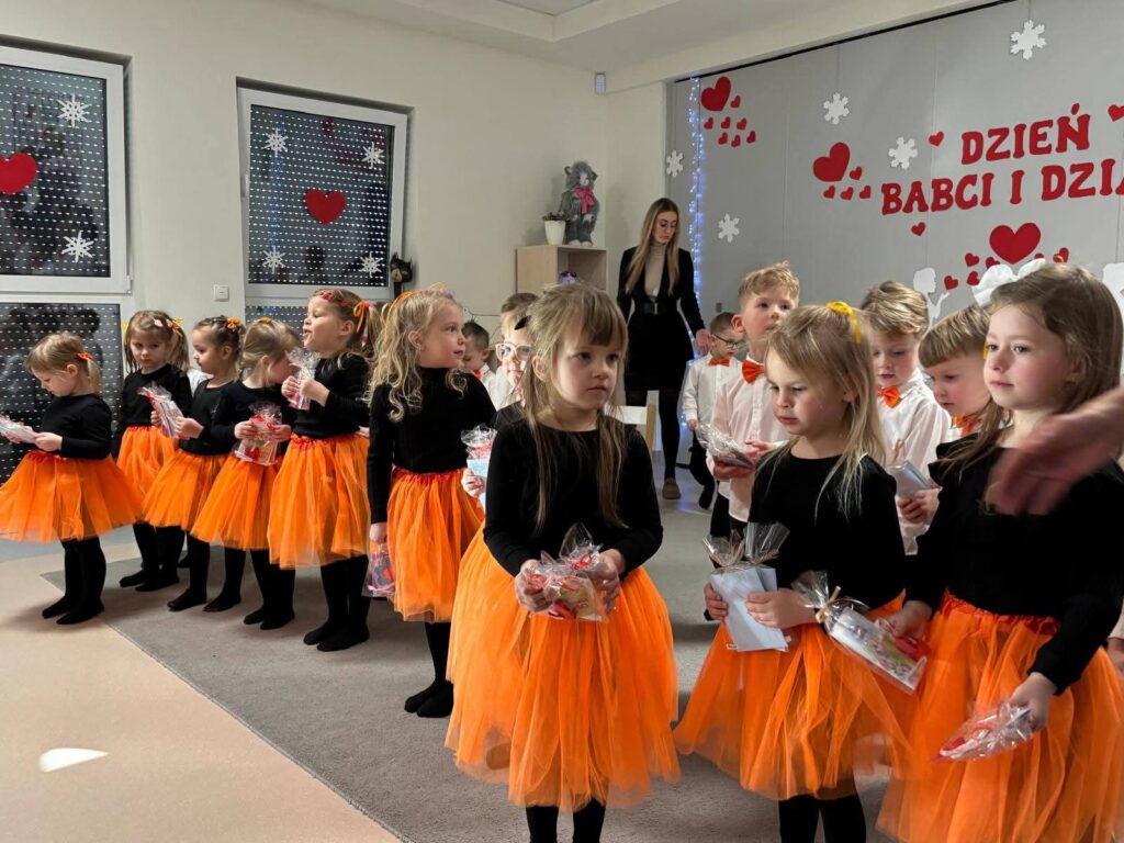 Duża grupa dzieci stoi na szarym dywanie, w rękach trzymają prezenty.