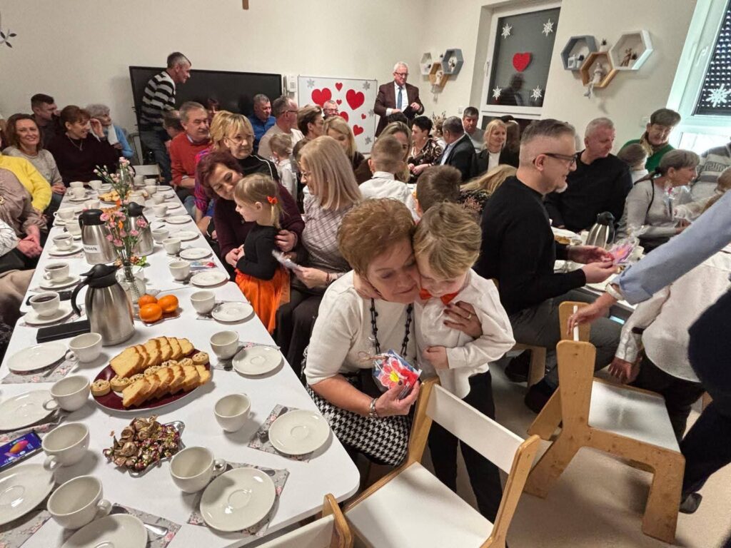 Dzieci składają życzenia babci i dziadkowi, którzy siedzą za białymi stołami, na których przygotowane jest ciasto.