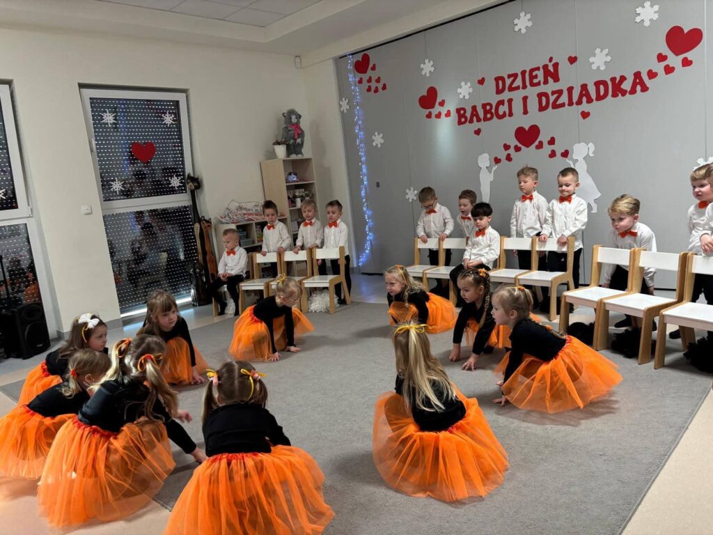Grupa dziewczynek ubrane na czarno w pomarańczowe spódniczki tańczą na szarym dywanie.