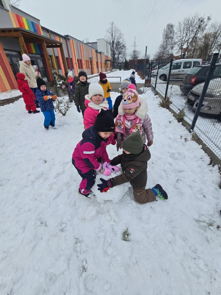 Dzieci ubrane w kolorowe kombinezony lepią duże kule śniegowe na bałwana.