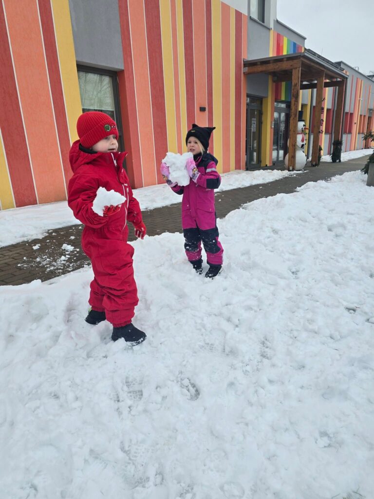 Dzieci ubrane w kolorowe kombinezony lepią duże kule śniegowe na bałwana.