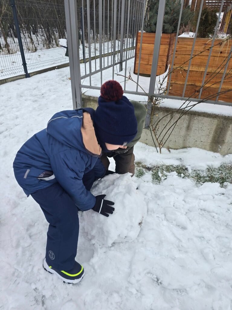 Dzieci ubrane w kolorowe kombinezony lepią duże kule śniegowe na bałwana.